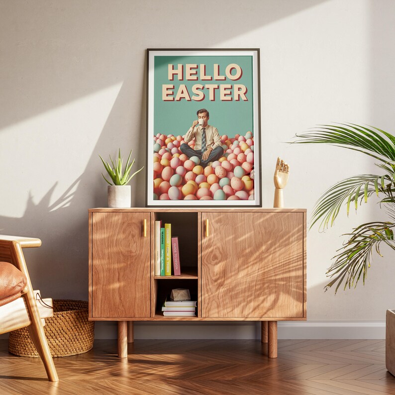 May include: A framed print with the text "HELLO EASTER". The image shows a person sitting in a pile of pastel Easter eggs. The print is displayed above a wooden cabinet with books and a small plant. The cabinet is made of wood.