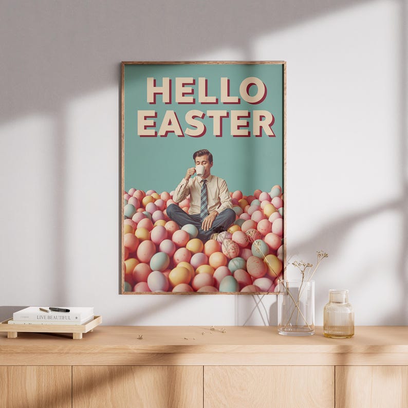 May include: A framed poster with the words "HELLO EASTER" in beige lettering. The image shows a man in a pile of pastel Easter eggs, drinking from a cup. The background is a teal colour.