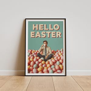 May include: A framed poster with the words "HELLO EASTER". The image shows a person sitting cross-legged in a pile of pastel-coloured Easter eggs, drinking from a white mug. The poster has a retro style.