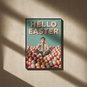 May include: A framed poster with the words "HELLO EASTER" in red and white. The image shows a man sitting in a pile of pastel Easter eggs, drinking from a cup. The background is a light teal colour.