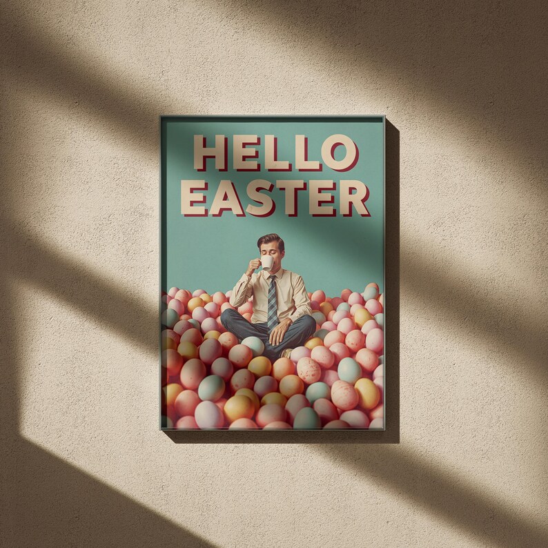 May include: A framed poster with the words "HELLO EASTER" in red and white. The image shows a man sitting in a pile of pastel Easter eggs, drinking from a cup. The background is a light teal colour.