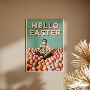 May include: A framed poster with the words "HELLO EASTER" in red and white letters. The poster shows a man sitting in a pile of colourful Easter eggs, drinking from a white mug. The background is a teal colour.