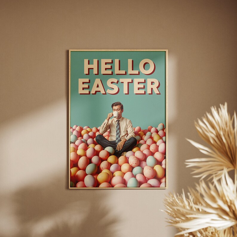 May include: A framed poster with the words "HELLO EASTER" in red and white letters. The poster shows a man sitting in a pile of colourful Easter eggs, drinking from a white mug. The background is a teal colour.