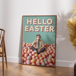 May include: A framed poster with the text "HELLO EASTER" in red and white. The poster shows a man seated in a pile of pastel-coloured eggs, holding a cup. The background is a light teal colour.