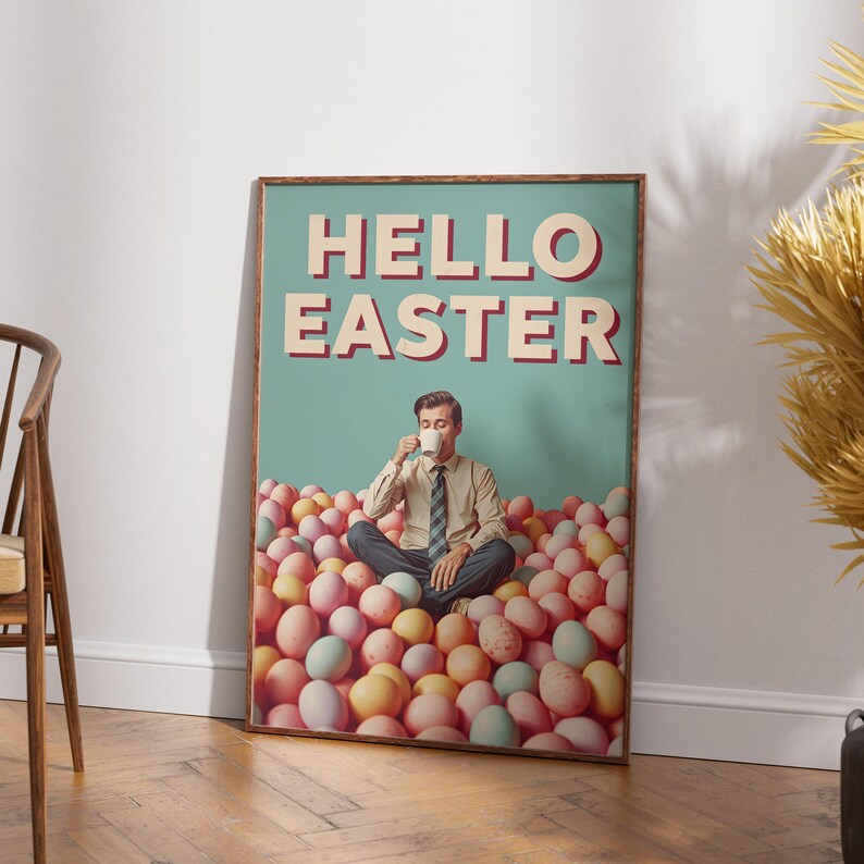 May include: A framed poster with the text "HELLO EASTER" in red and white. The poster shows a man seated in a pile of pastel-coloured eggs, holding a cup. The background is a light teal colour.