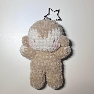 May include: A handmade crochet doll keychain in the shape of a small person. The doll is beige with white accents and pink cheeks. It has a silver star-shaped clasp for attaching to keys or bags.