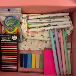May include: A pink box filled with stationery supplies. Includes colorful washi tape, sticky notes, a notepad, and pens in pastel colors. Also visible are several Mildliner double-ended highlighters.