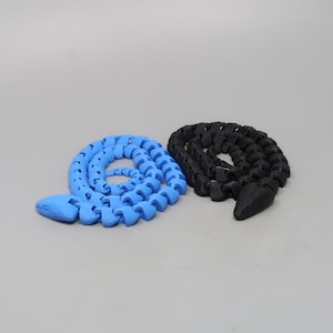 May include: Two articulated snake toys. One is light blue, the other is black. Each snake is coiled, with a segmented body and a pointed head. The toys are made of a solid material and are displayed against a neutral background.