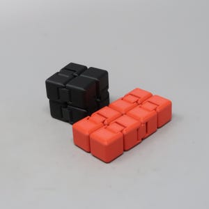 May include: Two fidget cubes are displayed against a gray background. One is black and cube-shaped, while the other is orange and rectangular. The orange cube is made of eight connected smaller cubes.