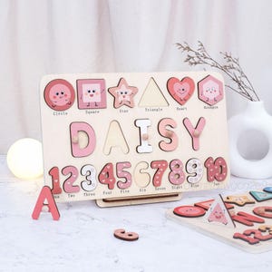 May include: Wooden educational puzzle featuring the name "DAISY" in pink letters, along with numbers 1-10 and various shapes. The puzzle includes a separate "A" piece and a partial view of a second puzzle. The shapes and numbers have cute faces.