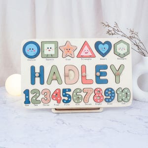 May include: Wooden puzzle board featuring the name "HADLEY" in colorful letters. The board includes shape and number cutouts with corresponding labels. Shapes include a circle, square, star, triangle, heart, and hexagon. Numbers 1-10 are also present.