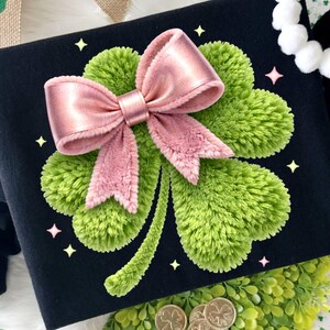 May include: Black sweatshirt featuring a large green shamrock with a pink bow. The shamrock is made of textured material. Gold coins and decorative shamrock accents are also visible, along with a banner with a shamrock.
