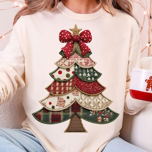 Op de afbeelding: Crèmekleurige sweatshirt met een kerstboomontwerp. De boom is samengesteld uit verschillende stoffen met patronen in rood, groen en crème, bekroond met een rode strik en gouden ster. Een rood-witte mok met een peperkoekmannetje is ook zichtbaar.