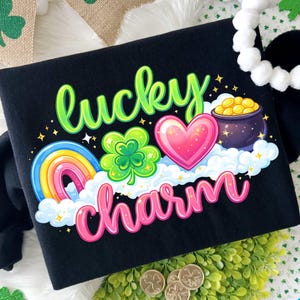Puede incluir: Camiseta negra con un diseño colorido de "Lucky Charm". El diseño incluye un arcoíris, un trébol, un corazón, una olla de oro y nubes. Banderas y acentos decorativos de tréboles rodean la camiseta.