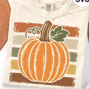 May include: A cream-colored t-shirt featuring a graphic of a large orange pumpkin with a green leaf and stem. The pumpkin is centered over five horizontal, distressed-texture stripes in shades of brown, beige, and olive green.