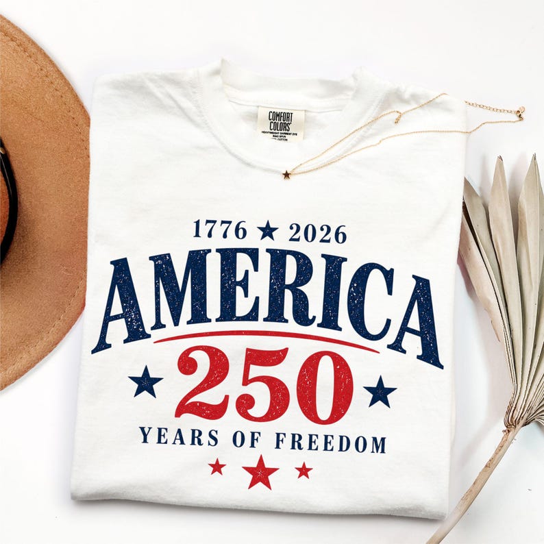 May include: White t-shirt with the text "AMERICA 250 YEARS OF FREEDOM" in blue and red. The shirt also features the years "1776 2026" and star graphics. A gold necklace with a star pendant is on the shirt.