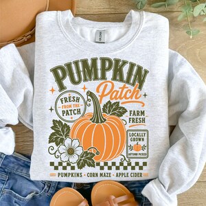 K&ouml;nnte beinhalten: Ein hellgraues Sweatshirt mit Rundhalsausschnitt und einem Grafikdruck mit dem Text PUMPKIN Patch, FRESH FROM THE PATCH, FARM FRESH, LOCALLY GROWN, PUMPKINS, CORN MAZE und APPLE CIDER. Das Design zeigt einen gro&szlig;en orangefarbenen K&uuml;rbis und Bl&auml;tter.