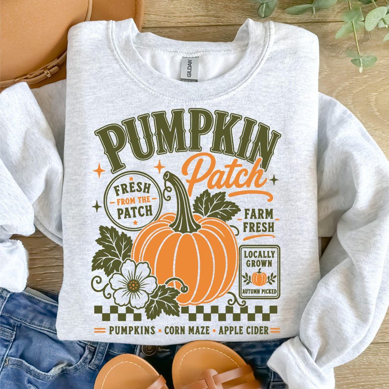 K&ouml;nnte beinhalten: Ein hellgraues Sweatshirt mit Rundhalsausschnitt und einem Grafikdruck mit dem Text PUMPKIN Patch, FRESH FROM THE PATCH, FARM FRESH, LOCALLY GROWN, PUMPKINS, CORN MAZE und APPLE CIDER. Das Design zeigt einen gro&szlig;en orangefarbenen K&uuml;rbis und Bl&auml;tter.