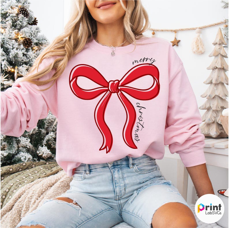 Puede incluir: Sudadera rosa claro con un gran dise&ntilde;o de lazo rojo y las palabras "merry christmas" en fuente cursiva. El lazo tiene un contorno blanco. Una prenda festiva para las fiestas.