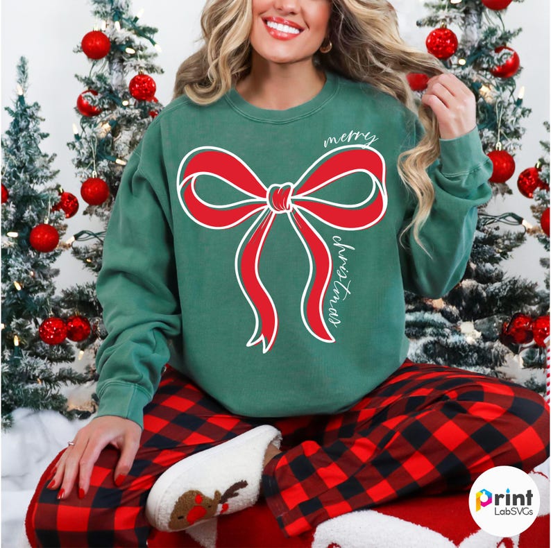 Puede incluir: Sudadera navide&ntilde;a verde con un gran dise&ntilde;o de lazo rojo y las palabras "merry" y "christmas" en escritura blanca. La persona lleva un pantal&oacute;n de pijama a cuadros rojos y negros. Decoraciones de &aacute;rbol de Navidad en el fondo.