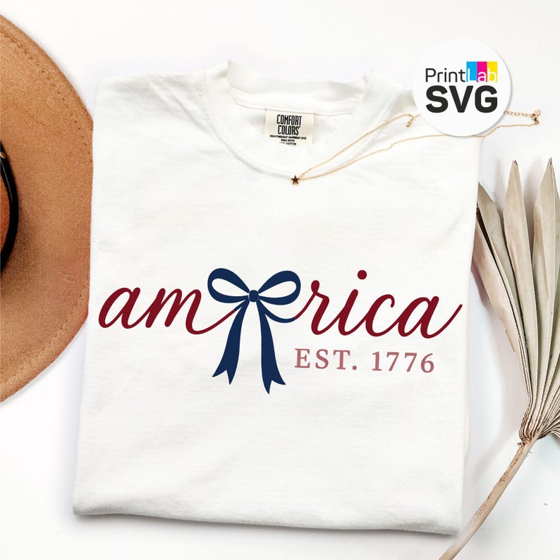 K&ouml;nnte beinhalten: Ein wei&szlig;es T-Shirt mit dem Wort "America" in Schreibschrift, mit einer marineblauen Schleife und dem Text "EST. 1776" in Rot. Das Shirt hat ein Comfort Colors-Etikett und eine goldene Halskette mit Sternanh&auml;nger.