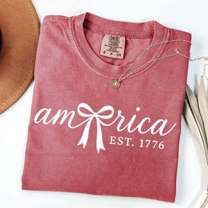K&ouml;nnte beinhalten: Ein staubrosa T-Shirt mit dem Wort "America" in wei&szlig;er Schrift, mit einer Bogen-Grafik. Der Text "EST. 1776" steht unter dem Wort. Eine silberne Halskette mit Anh&auml;nger befindet sich auf dem Shirt.