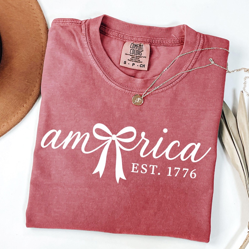 K&ouml;nnte beinhalten: Ein staubrosa T-Shirt mit dem Wort "America" in wei&szlig;er Schrift, mit einer Bogen-Grafik. Der Text "EST. 1776" steht unter dem Wort. Eine silberne Halskette mit Anh&auml;nger befindet sich auf dem Shirt.
