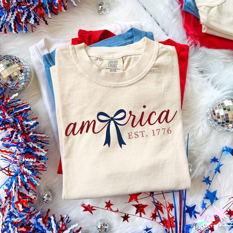 K&ouml;nnte beinhalten: Cremefarbenes T-Shirt mit dem Wort "america" in burgunderfarbener Schrift und einer blauen Schleifen-Grafik. Der Text "EST. 1776" steht darunter. Das Shirt ist Teil eines Stapels roter, wei&szlig;er und blauer Shirts.