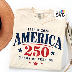May include: Cream-colored t-shirt with the text "AMERICA 250 YEARS OF FREEDOM" in navy blue and red. The design includes stars and the years 1776 and 2026. The shirt is made of Comfort Colors material.