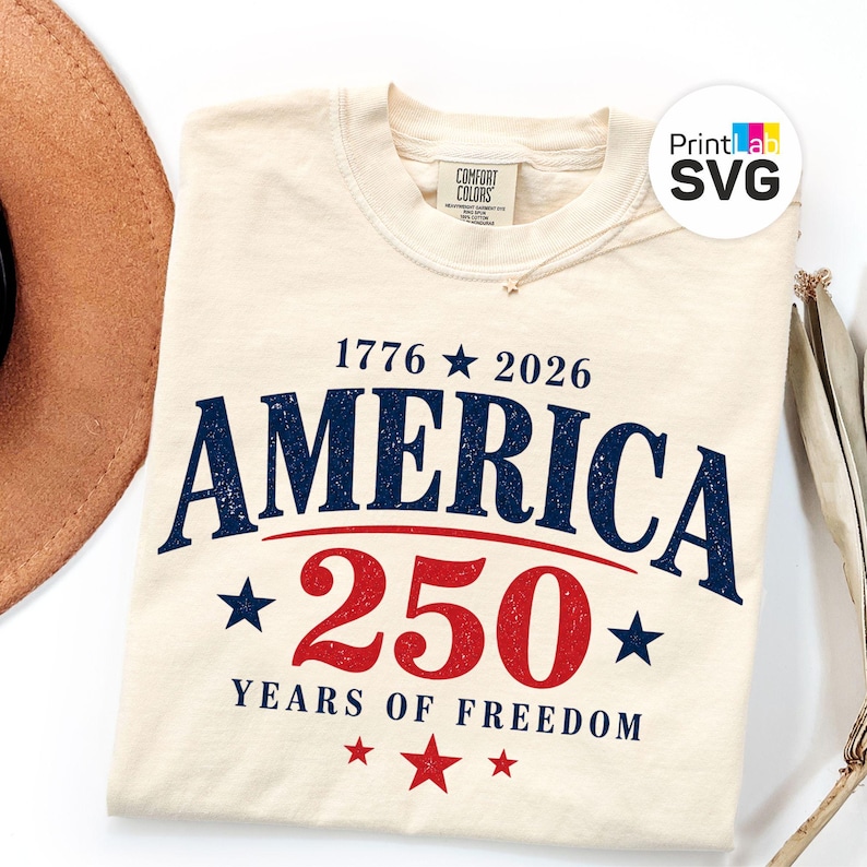 May include: Cream-colored t-shirt with the text "AMERICA 250 YEARS OF FREEDOM" in navy blue and red. The design includes stars and the years 1776 and 2026. The shirt is made of Comfort Colors material.