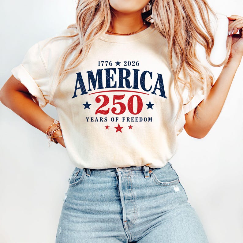 May include: Cream-colored t-shirt with the text "AMERICA 250 YEARS OF FREEDOM" in navy blue and red. The design includes stars and the years 1776 and 2026. The shirt is paired with blue jeans.