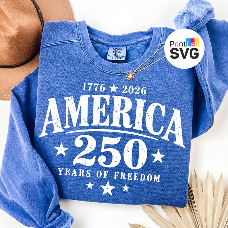 May include: A blue sweatshirt with the text "1776 2026 AMERICA 250 YEARS OF FREEDOM" in white. The sweatshirt has a crew neck and long sleeves. A gold necklace is draped on the sweatshirt.