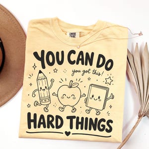 Op de afbeelding: Een lichtgeel t-shirt met een zwarte grafische print van een lachend potlood, een appel en een boek, met de tekst "YOU CAN DO HARD THINGS" en "you got this!" en kleine sterretjes. Het shirt heeft een Comfort Colors label in de nek.