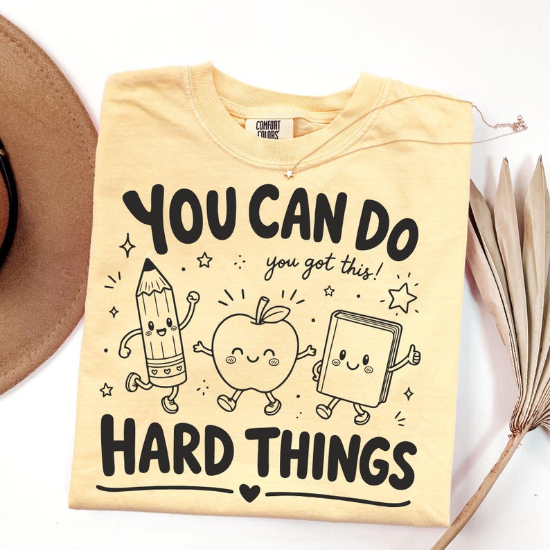 Op de afbeelding: Een lichtgeel t-shirt met een zwarte grafische print van een lachend potlood, een appel en een boek, met de tekst "YOU CAN DO HARD THINGS" en "you got this!" en kleine sterretjes. Het shirt heeft een Comfort Colors label in de nek.