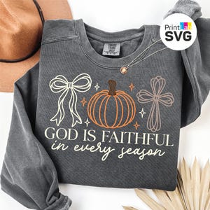 Puede incluir: Una sudadera gris carbón de cuello redondo con un estampado gráfico que muestra un lazo blanco, una calabaza naranja y una cruz de color canela. El texto "GOD IS FAITHFUL in every season" aparece en una mezcla de fuentes de bloque y cursiva.
