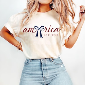 K&ouml;nnte beinhalten: Cremefarbenes T-Shirt mit dem Wort "America" in Schreibschrift, mit einer marineblauen Schleife und dem Text "EST. 1776". Das Shirt wird mit hellblauen Jeans getragen.
