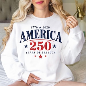 May include: White sweatshirt with the text "AMERICA 250 YEARS OF FREEDOM" in navy blue and red. The design includes stars and the years 1776 and 2026. A casual, patriotic apparel item.