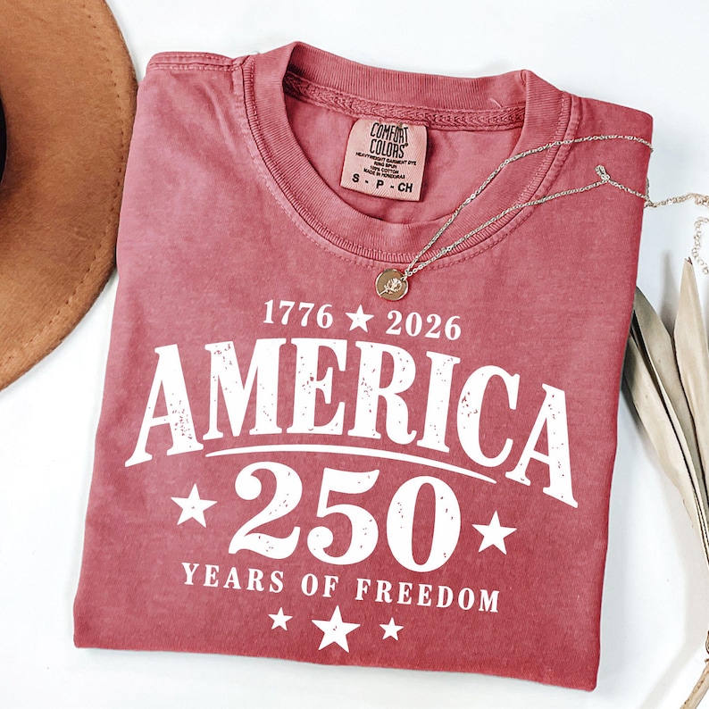 May include: A dusty rose-colored t-shirt with the text "AMERICA 250 YEARS OF FREEDOM" in white. The shirt has a crew neck and short sleeves. A silver necklace with a pendant is draped across the shirt.