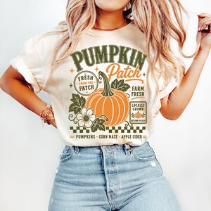 K&ouml;nnte beinhalten: Ein cremefarbenes T-Shirt mit einem Grafikdruck, der einen orangefarbenen K&uuml;rbis, gr&uuml;ne Bl&auml;tter und den Text PUMPKIN Patch, FRESH FROM THE PATCH, FARM FRESH, LOCALLY GROWN, AUTUMN PICKED, PUMPKINS, CORN MAZE und APPLE CIDER zeigt.