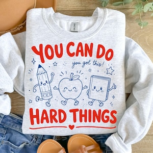 Op de afbeelding: Een lichtgrijs sweatshirt met ronde hals en een grafische print met de rode tekst YOU CAN DO en HARD THINGS, en de blauwe tekst you got this! Het ontwerp bevat cartoonillustraties van een potlood, een appel en een boek met gezichtjes.