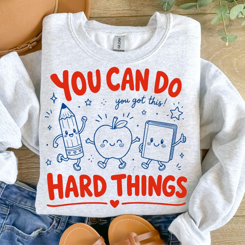 Op de afbeelding: Een lichtgrijs sweatshirt met ronde hals en een grafische print met de rode tekst YOU CAN DO en HARD THINGS, en de blauwe tekst you got this! Het ontwerp bevat cartoonillustraties van een potlood, een appel en een boek met gezichtjes.