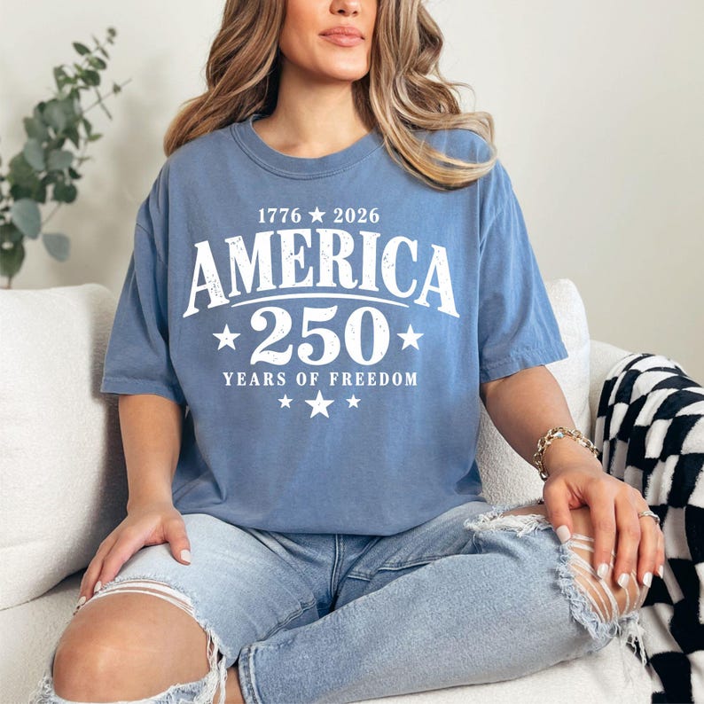 May include: A blue t-shirt with the text "AMERICA 250 YEARS OF FREEDOM" in white. The shirt also has the years "1776 2026" above the word "AMERICA". The shirt is a crew neck and has short sleeves.