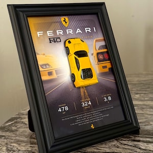 May include: A black framed display featuring a yellow Ferrari F40 model car mounted on a printed background. The print includes the Ferrari logo, the text FERRARI F40, and performance statistics: 478 HP, 324 KM/H top speed, and 3.8 seconds 0-100 KM/H.