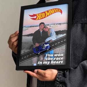 May include: A black framed shadow box containing a photograph of two people, a Hot Wheels logo at the top, a diagonal road graphic, a blue toy car attached to the surface, and the text "You won the race in my heart" printed in white.