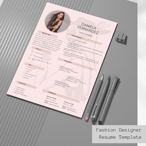 May include: A pink resume template for a fashion designer, featuring a photo, contact details, skills, and work experience. The resume includes the text "Fashion Designer Resume Template" at the bottom. Two pens, a pencil, and a pencil sharpener are on the gray surface.