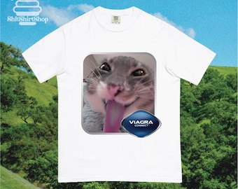 May include: White t-shirt featuring a gray cat with its tongue out, inside a square frame. Below the cat is a blue and silver diamond-shaped logo with the text "VIAGRA CONNECT". The shirt also has the logo "ShitShirtShop" in the upper left corner.
