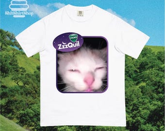 Könnte beinhalten: Weißes T-Shirt mit einer Grafik einer schläfrigen Katze in einem lila Rahmen, mit dem Vicks ZzzQuil Logo. Das Shirt hat eine lockere Passform. Der Hintergrund zeigt einen grünen Hügel und blauen Himmel.