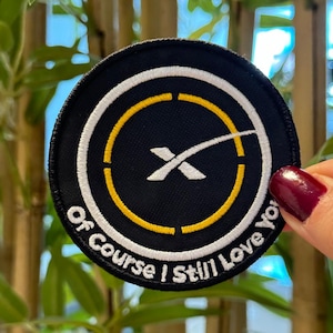 May include: A round black patch with a white and yellow design, featuring a stylized 'X' and the text 'Of Course I Still Love You'. The patch is likely an embroidered emblem.