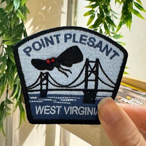 Op de afbeelding: Geborduurde patch met de tekst "POINT PLEASANT" en "WEST VIRGINIA". De patch toont een brug en een zwart wezen met rode ogen. Het heeft een lichtblauwe achtergrond en een zwarte rand.