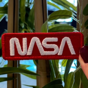 May include: A rectangular red patch with the white embroidered letters "NASA". The patch is a rectangle with rounded corners. The patch is held in front of a background of green leaves and bamboo stalks.